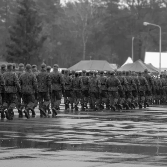Schwarz Weiss Fotografie. Soldat:innen maschieren im Gleichschritt diagonal weg vom Betrachter. Sie tragen Uniform aber keine Kampfausrüstung